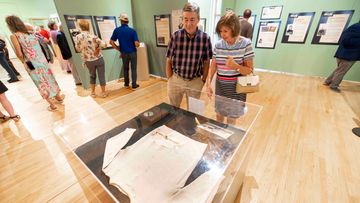  A man and woman look down at a display case containing a white sweatshirt in the middle of a gallery. The man wears a red and black plaid shirt, and the woman wears a blue and white striped shirt and a black and white striped skirt. A group of people are