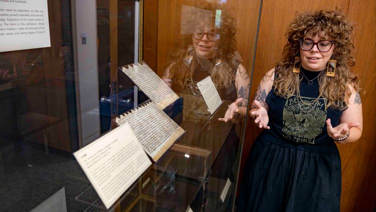  A person with curly brown hair and glasses stands next to a display case, gesturing towards open manuscripts inside. They are wearing a black top with a gold design and large earrings that look like guillotines . Their reflection is visible in the glass 